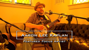 Aaron Lee Kaplan playing a resonator guitar into a microphone with text reading Aaron Lee Kaplan, Featured Focus Artist